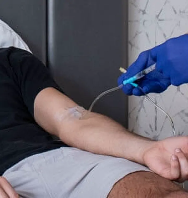 Close-up of an IV line taped to a patient's forearm, the tubing held by a provider's blue-gloved hand