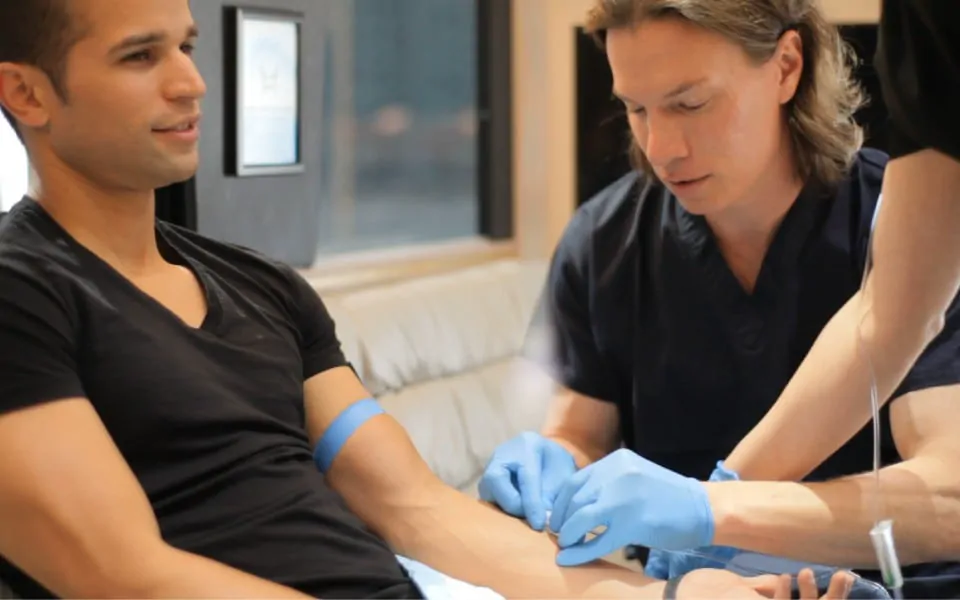 A Hangover Heaven clinician in navy scrubs and blue gloves places an IV in a smiling patient's arm