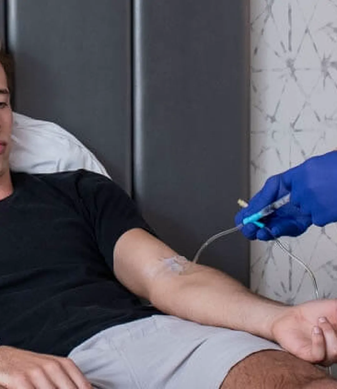 A patient reclines with an IV line in his forearm while a provider in blue gloves checks the tubing