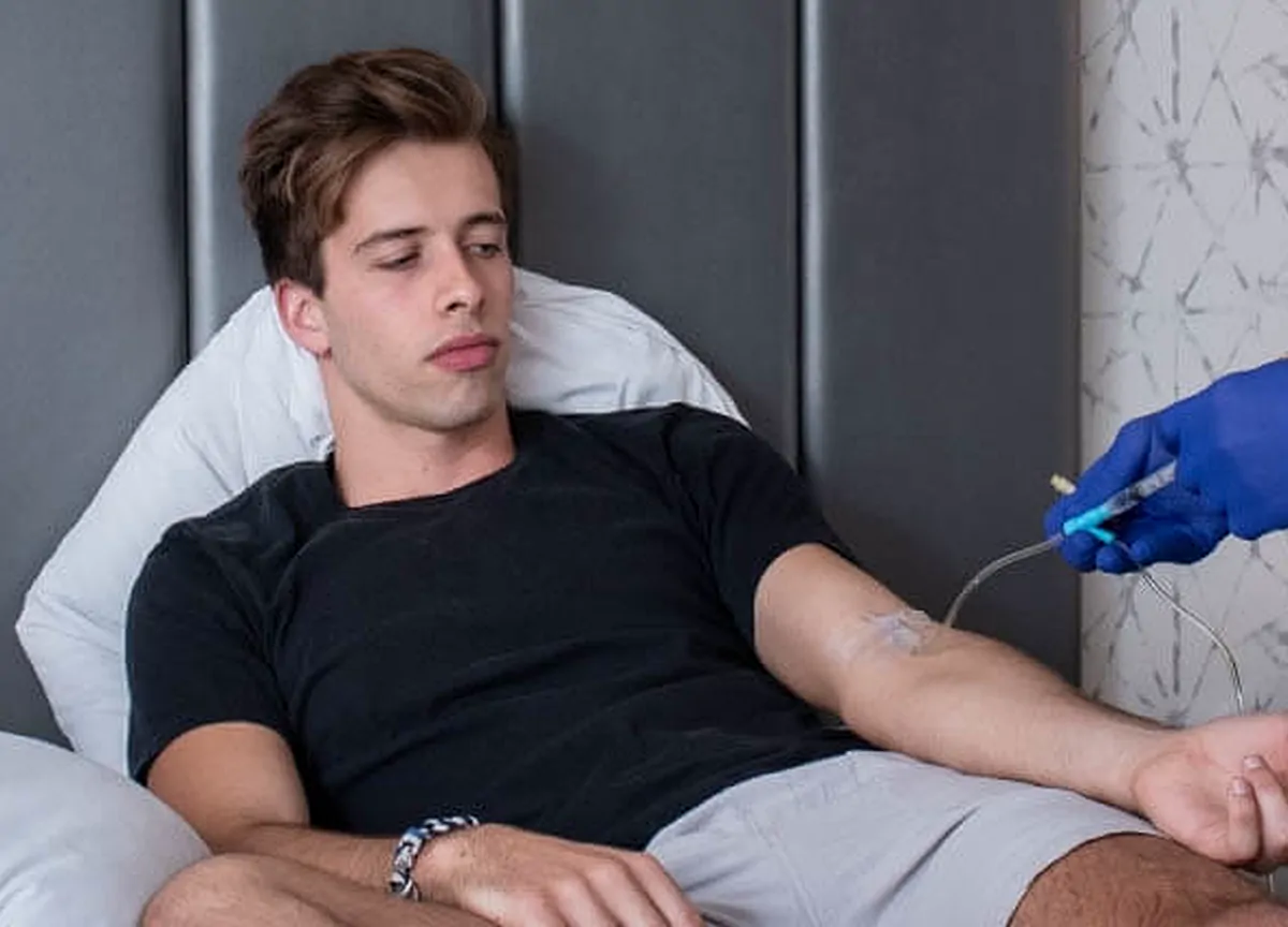 A young man in a black T-shirt reclines in a treatment chair while a provider in blue gloves adjusts the IV line in his arm